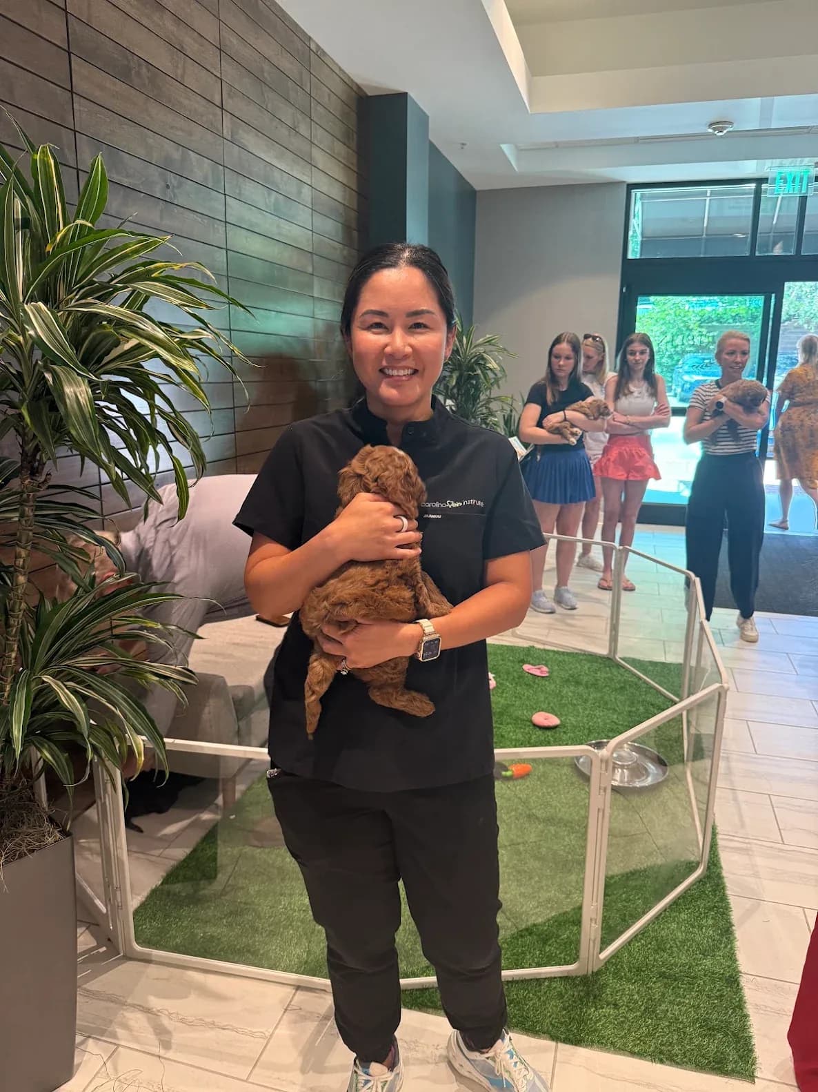 A Carolina Vein Institute staff member holding an apricot Australian Labradoodle puppy beside the play pen, with colleagues waiting their turn behind her